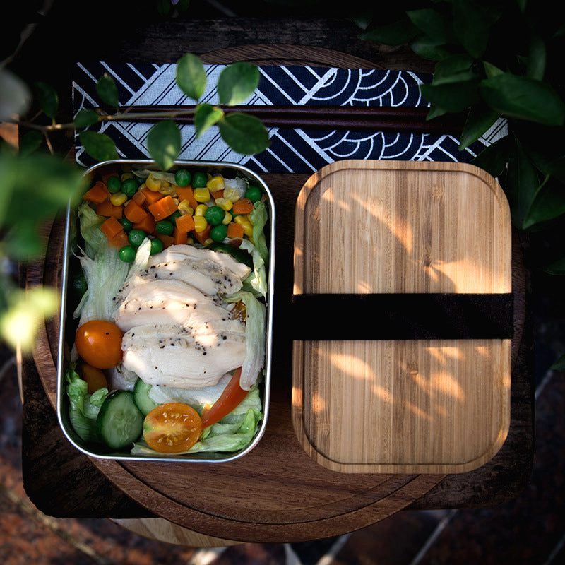 Bento en inox ouvert garni de poulet et légumes, couvercle en bambou à côté, vue de dessus