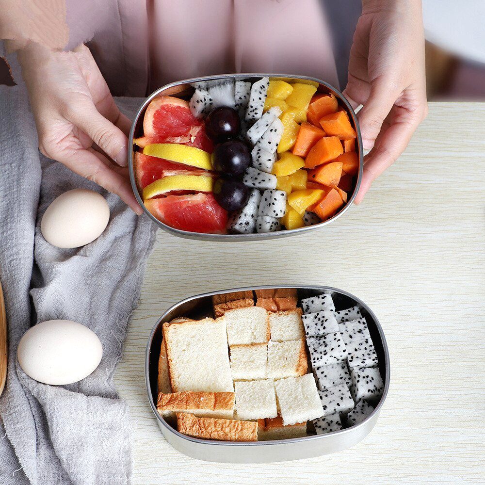 Deux bentos ovales en inox garnis de fruits et de pain, tenus en main